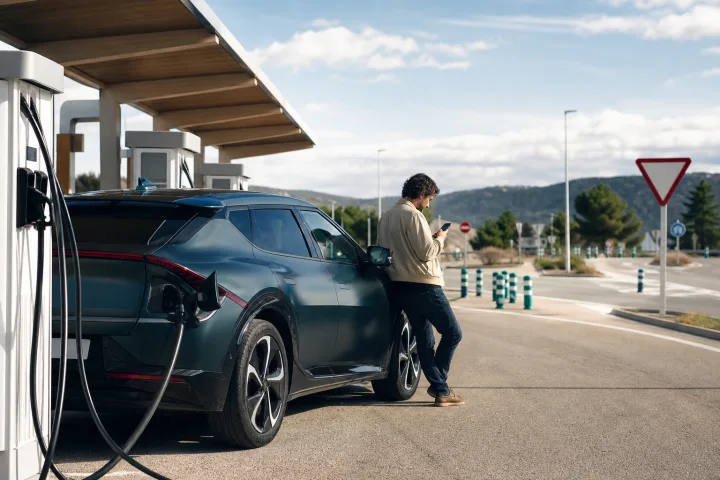 Elektroauto neben öffentlichen Ladepunkten an einer spanischen Raststätte, während eine Person die Route auf dem Smartphone prüft.