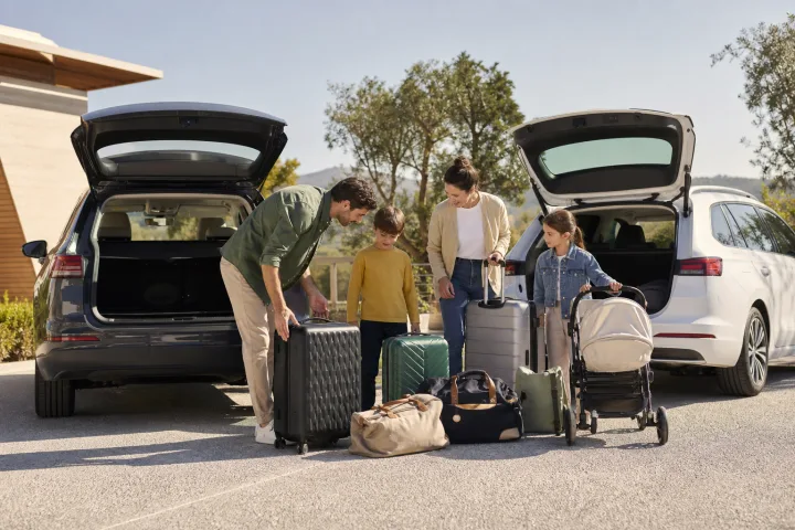 Família a comparar o espaço de carga de dois carros, com malas e um carrinho de bebé junto deles.